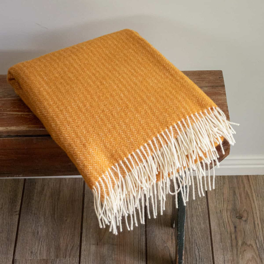 Orange throw blanket with white fringe on a wooden bench against a neutral wall.