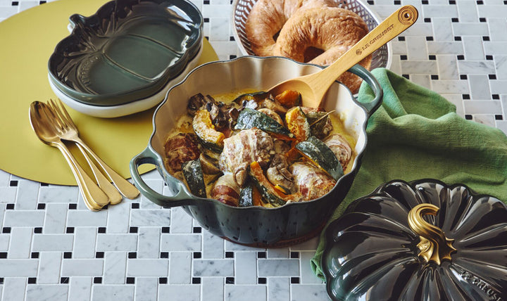 Dish of food with a wooden spoon on a tiled table, surrounded by kitchen items.