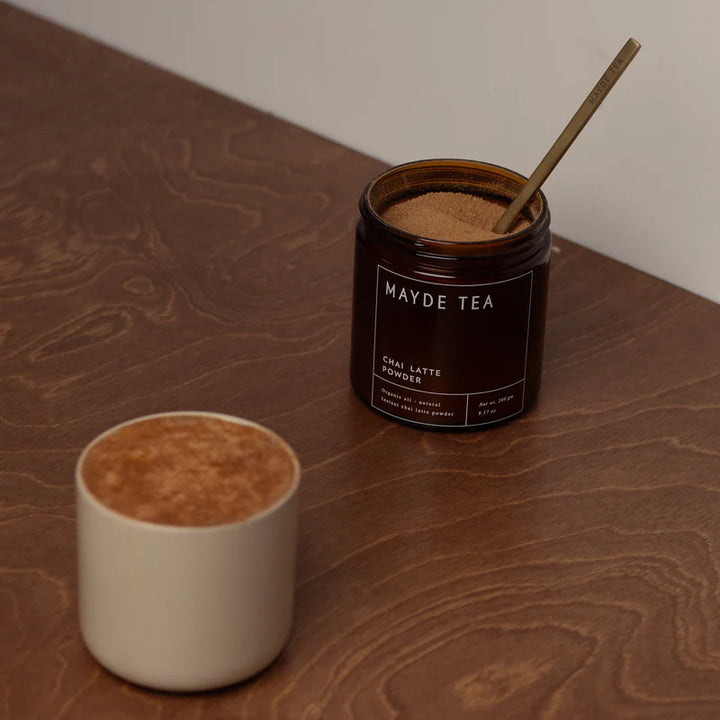 Mayde Tea Chai Latte Powder jar with a wooden spoon and a cup of chai on a wooden surface.
