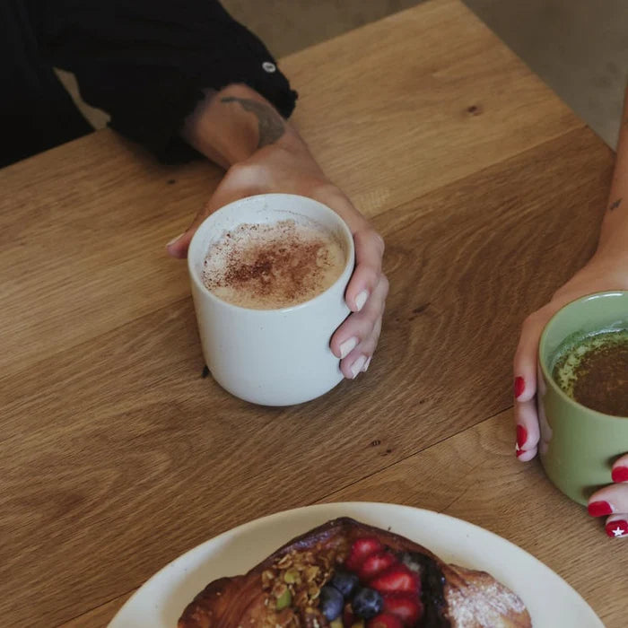 Two people holding coffee cups on a wooden table with a plate of food.