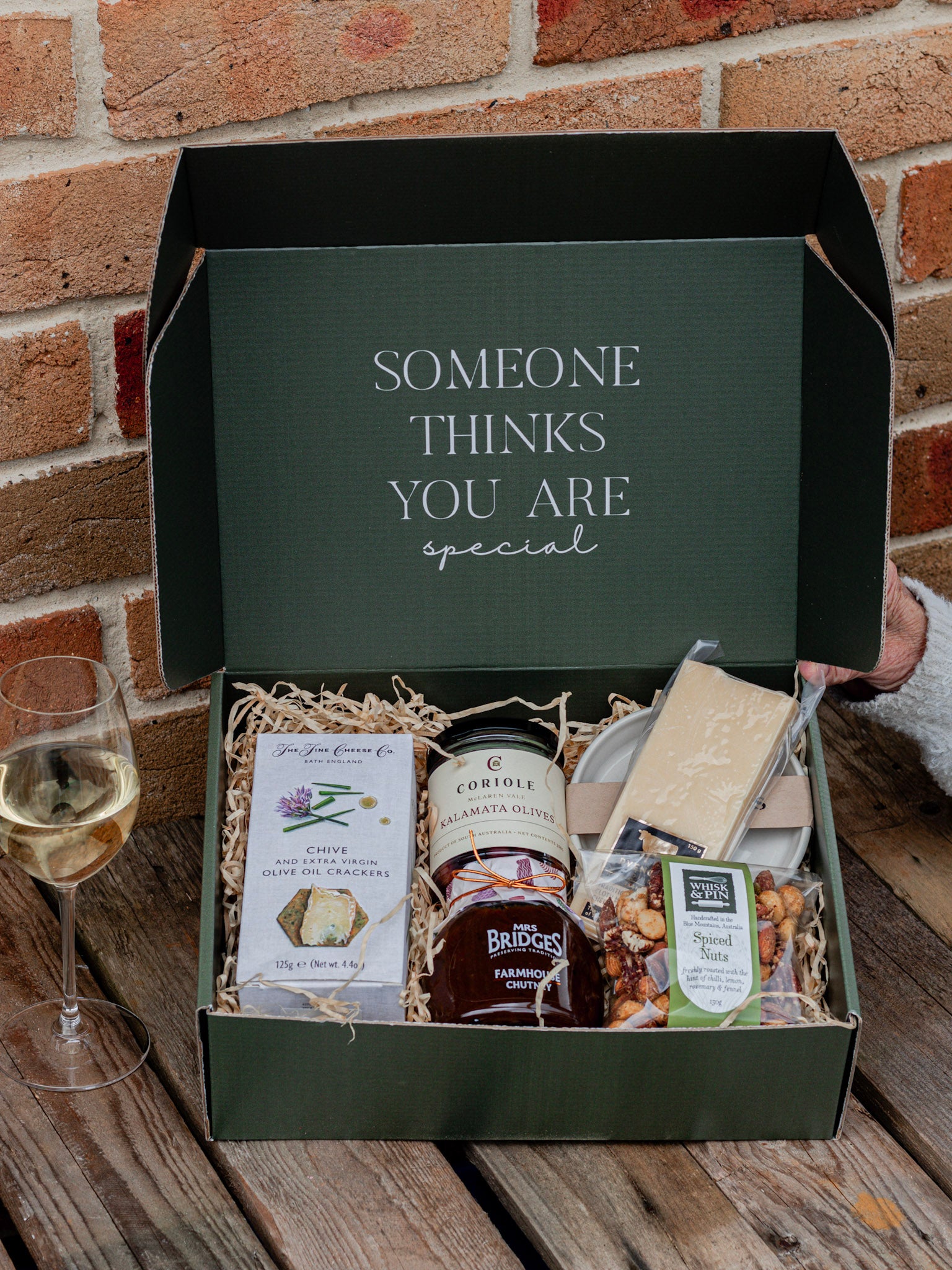 Gift box with products and a glass of wine on a wooden surface with a brick wall background.