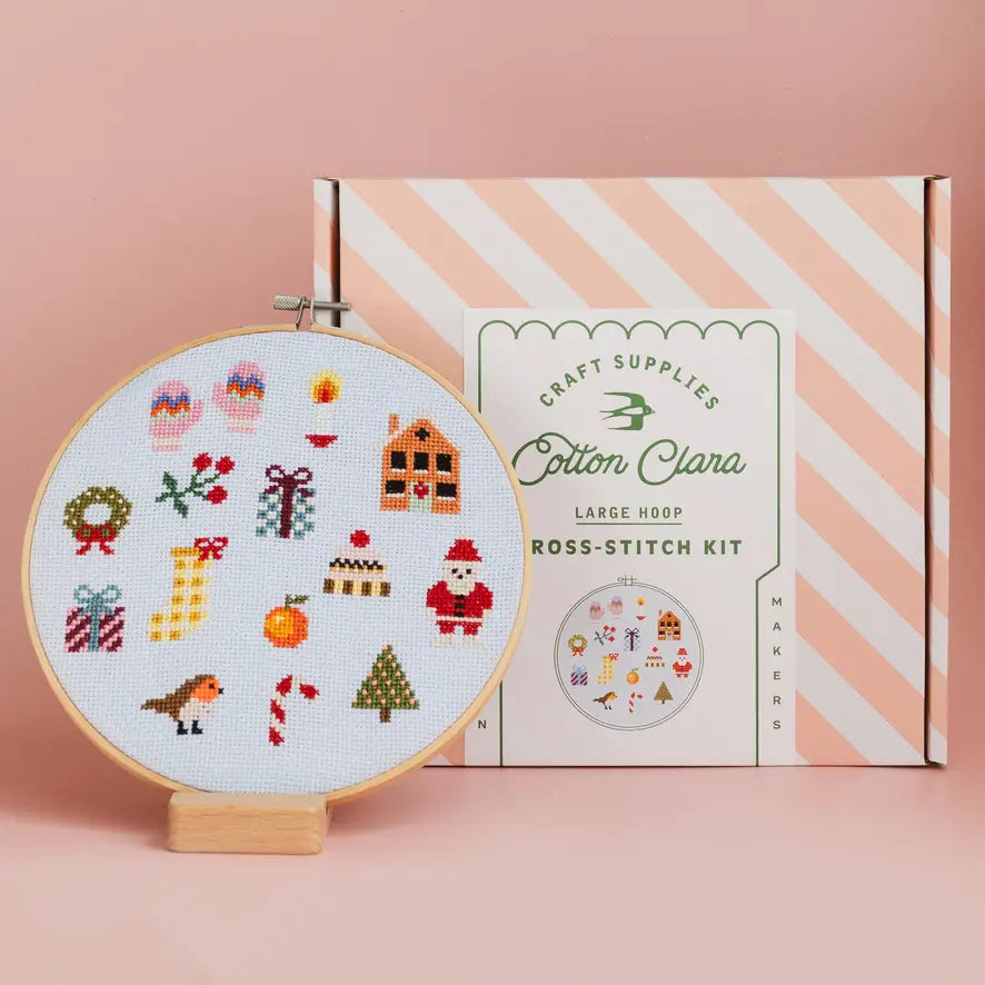 Cross-stitch hoop with festive designs and Cotton Clara stitching kit box on a pink background