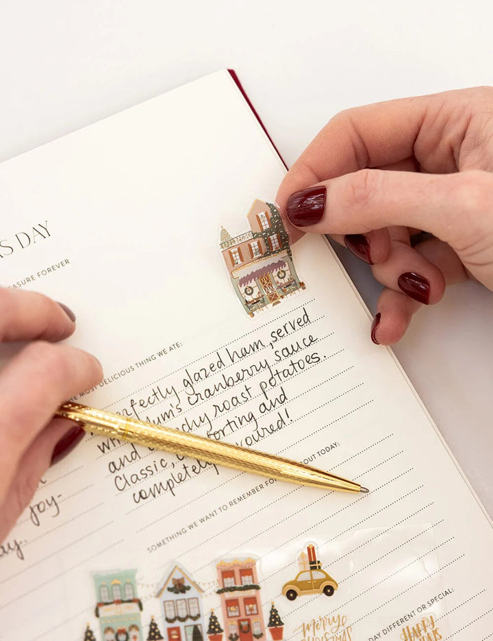 Person using a gold pen to write in a notebook with decorative stickers on a white surface