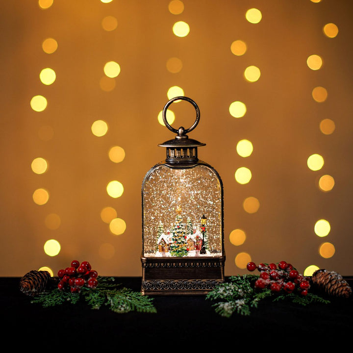Decorative lantern with Christmas scene inside, surrounded by festive decorations against a warm bokeh background.