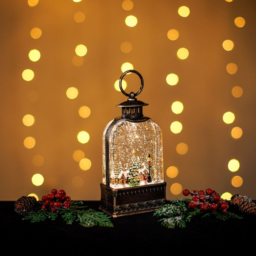 Decorative lantern with a Christmas scene inside, surrounded by festive decorations and lights on a blurred background.