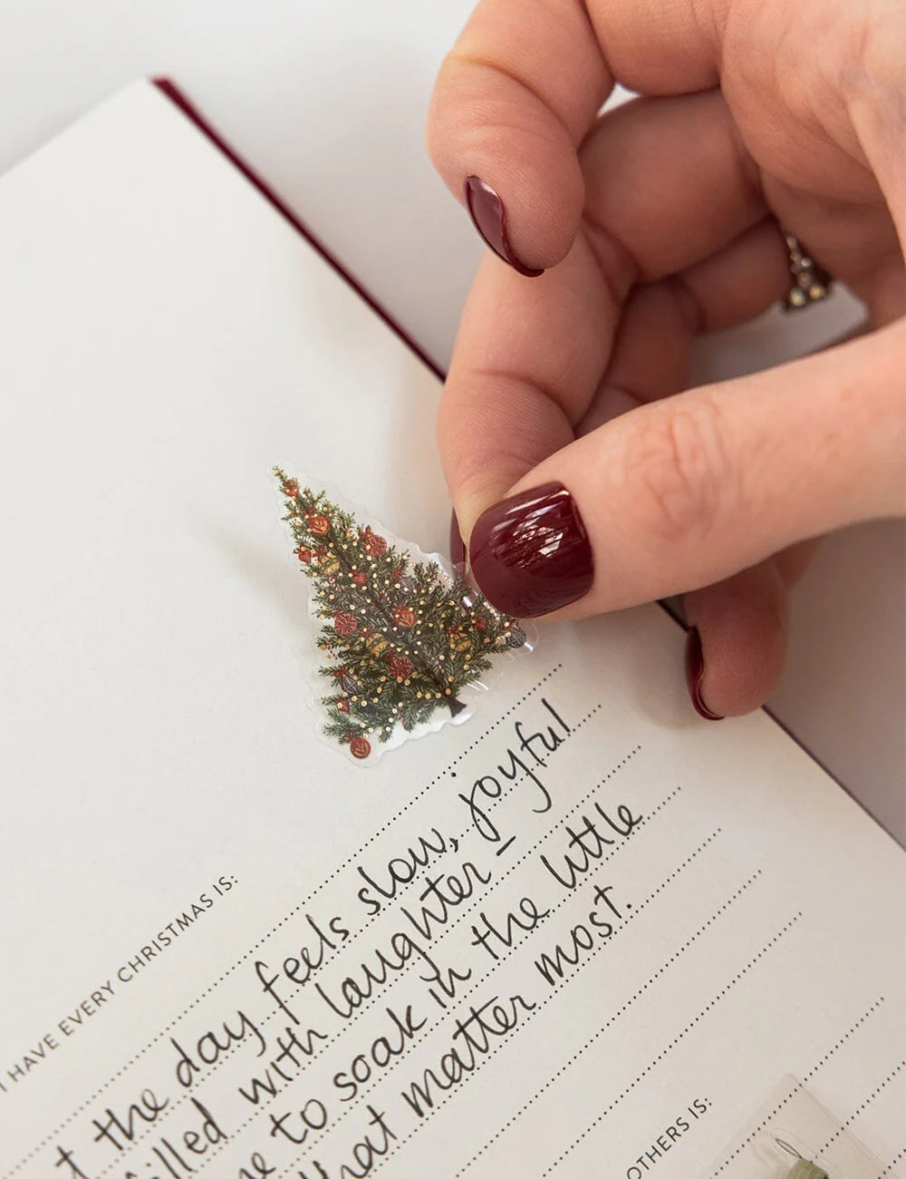 Hand with red nail polish holding a small Christmas tree decoration over a handwritten note.