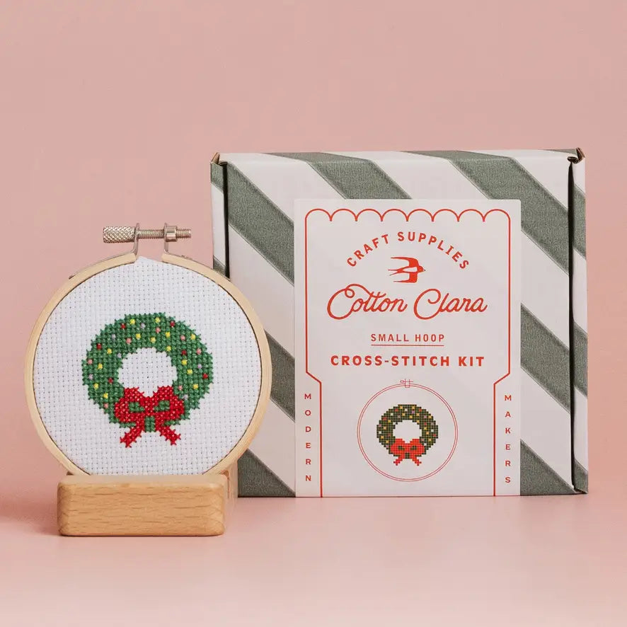 Cross-stitch kit with a completed wreath design and Cotton Clara packaging on a pink background