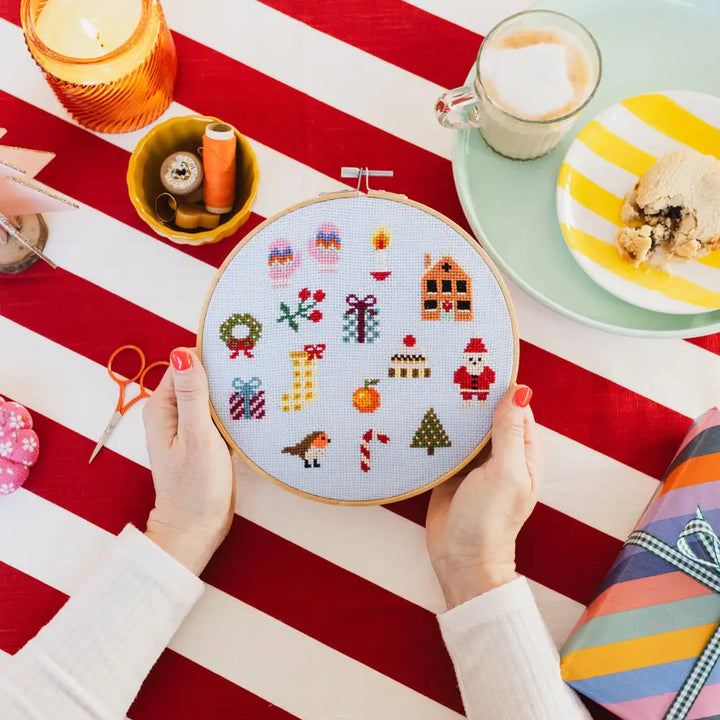 Person holding a cross-stitched hoop with festive designs on a table with a striped tablecloth.