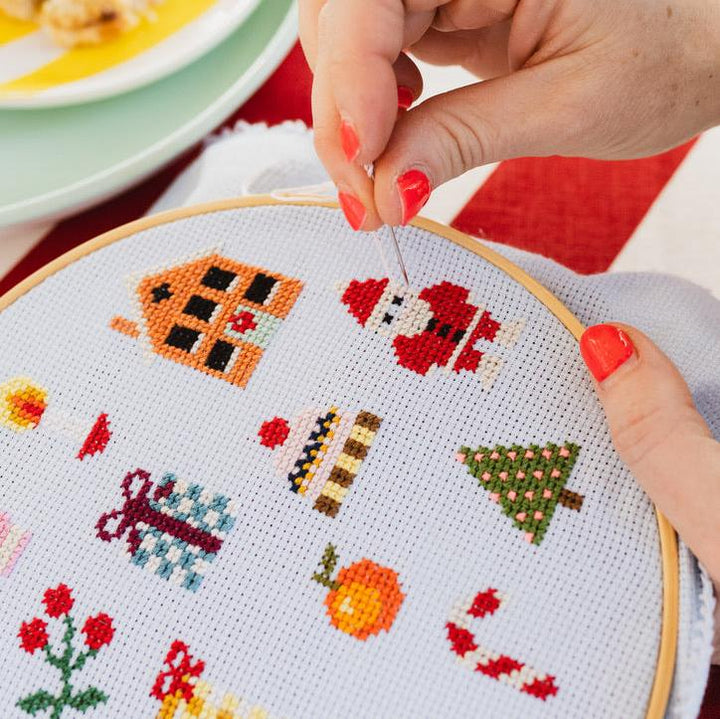 Cross-stitching project with festive designs on a white fabric, held by a person with red nail polish.