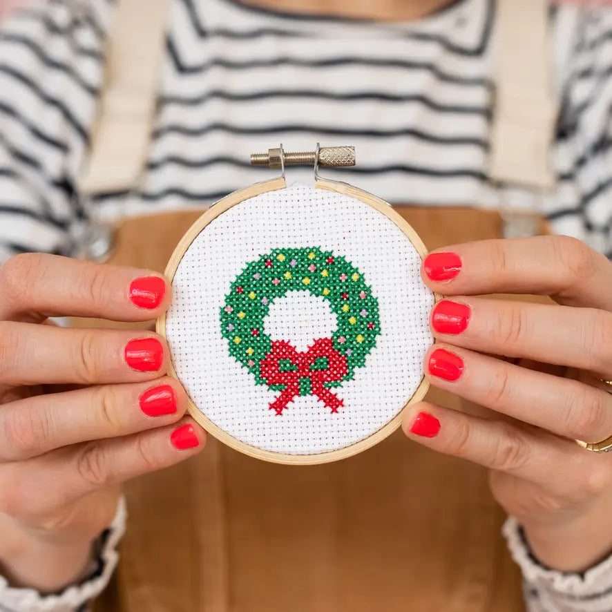 Person holding a cross-stitched Christmas wreath with red bow in a wooden hoop.