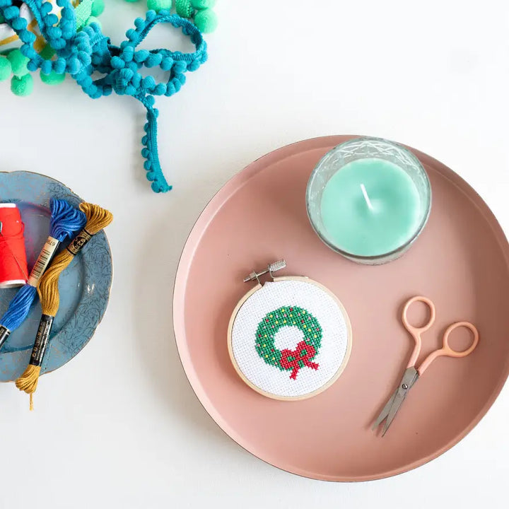 Cross-stitched wreath on a pink plate with scissors and thread, on a white background