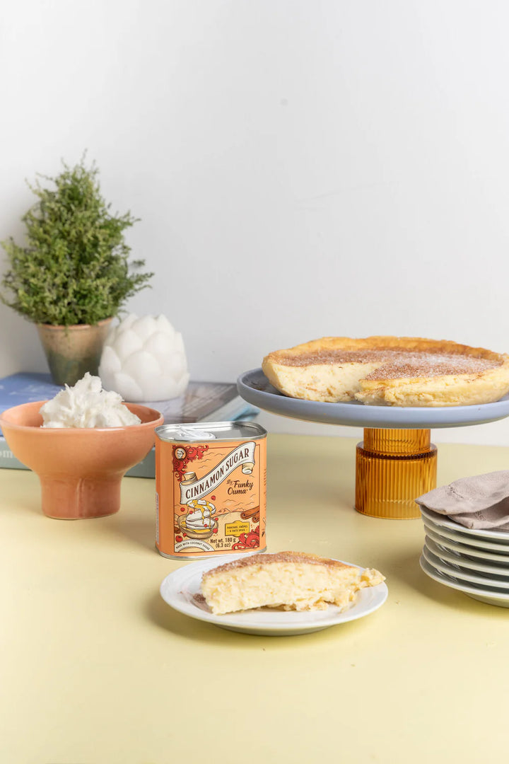 Cheesecake on a stand with a tin of cinnamon sugar and a slice on a plate on a yellow table 