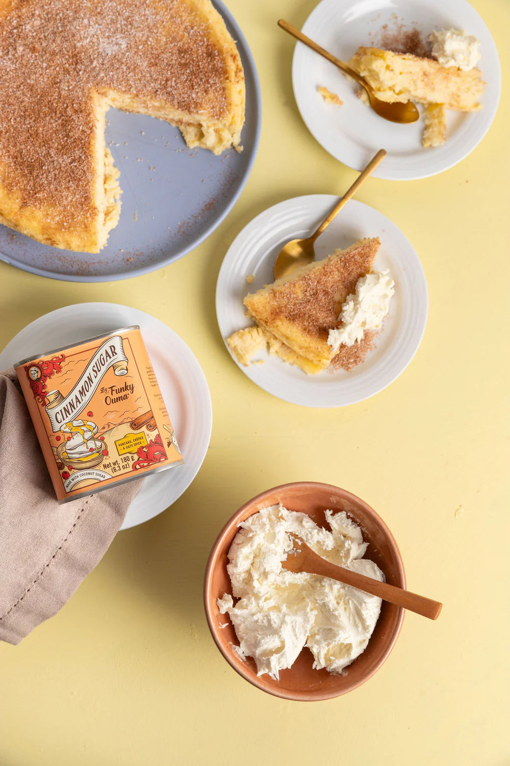 Sliced cake with whipped cream, cinnamon sugar tin, and bowl of whipped cream on a yellow background