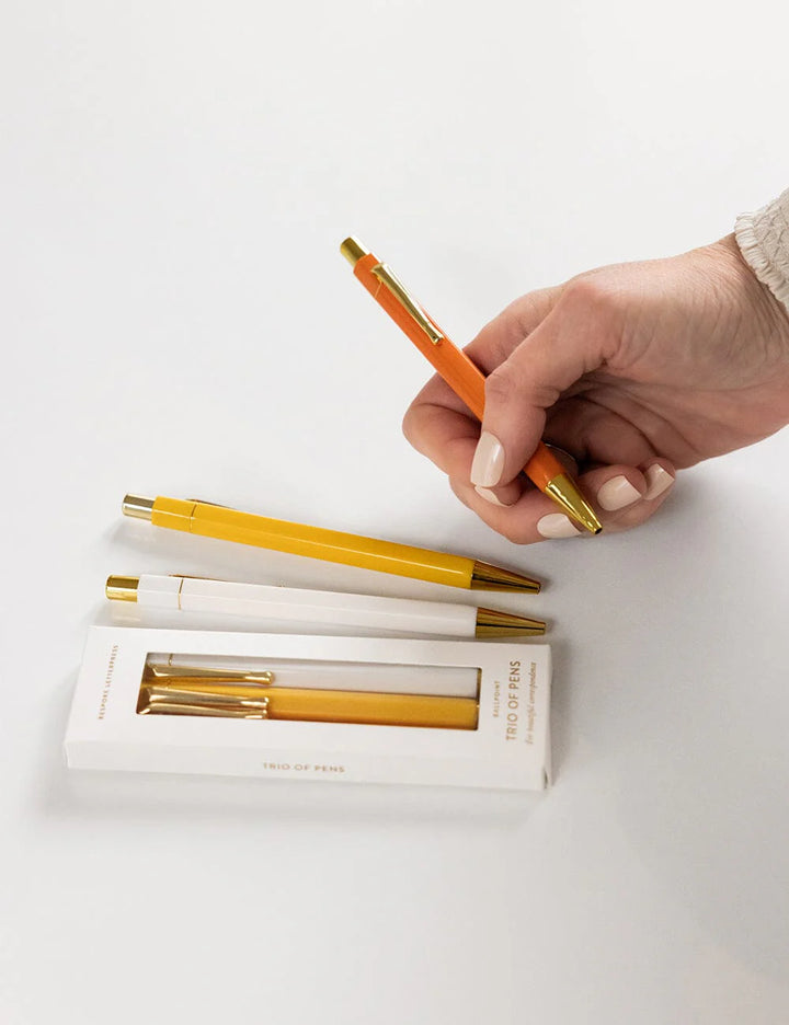 Hand holding an orange pen with two other pens and their packaging on a white background