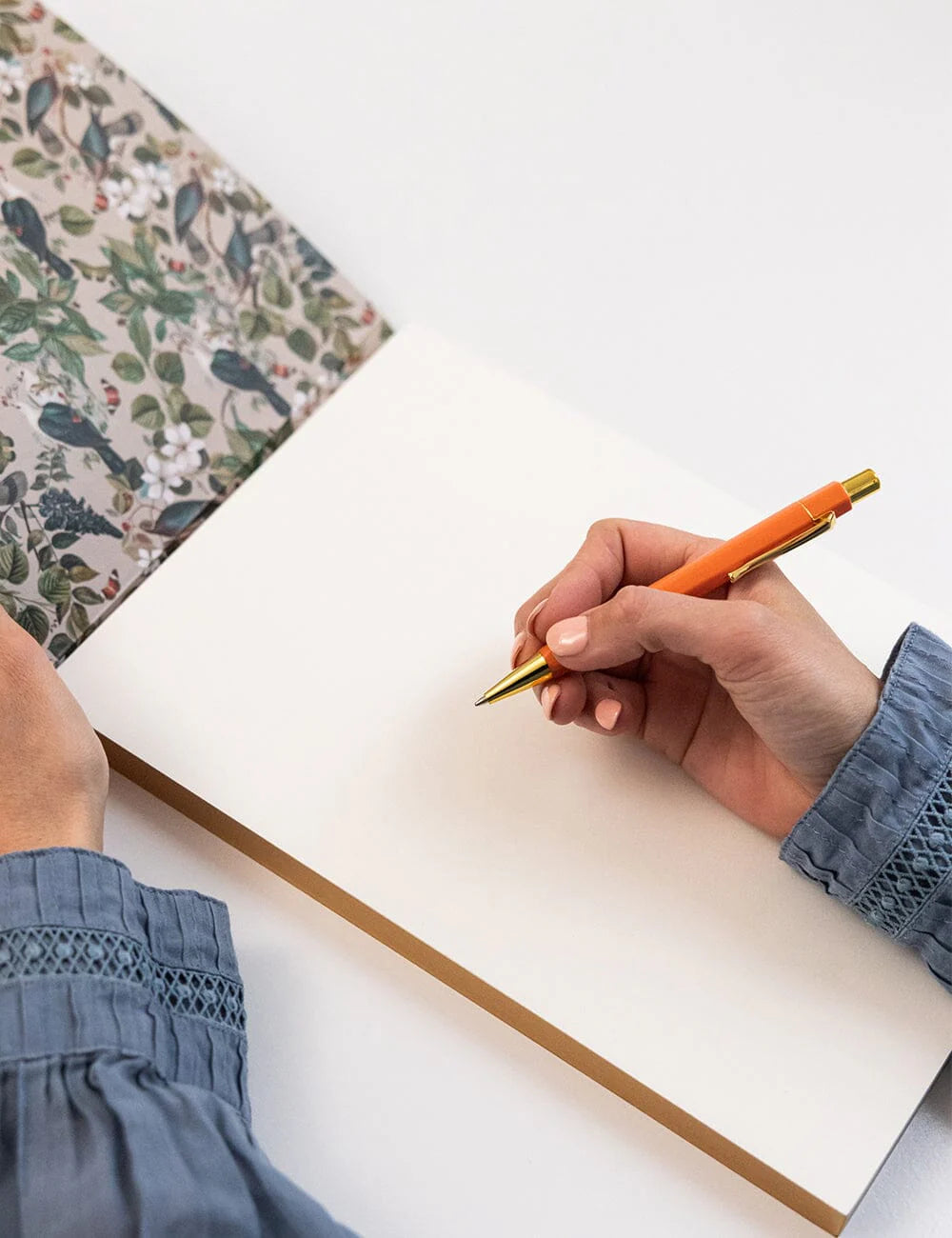 Hand holding a pen over an open notebook with a floral notebook in the background