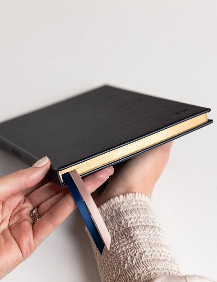 Hand holding a black leather-bound book against a light background
