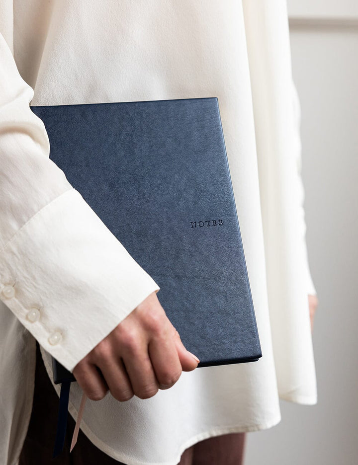 Person holding a blue notebook with 'Note' embossed on it, wearing a white coat.