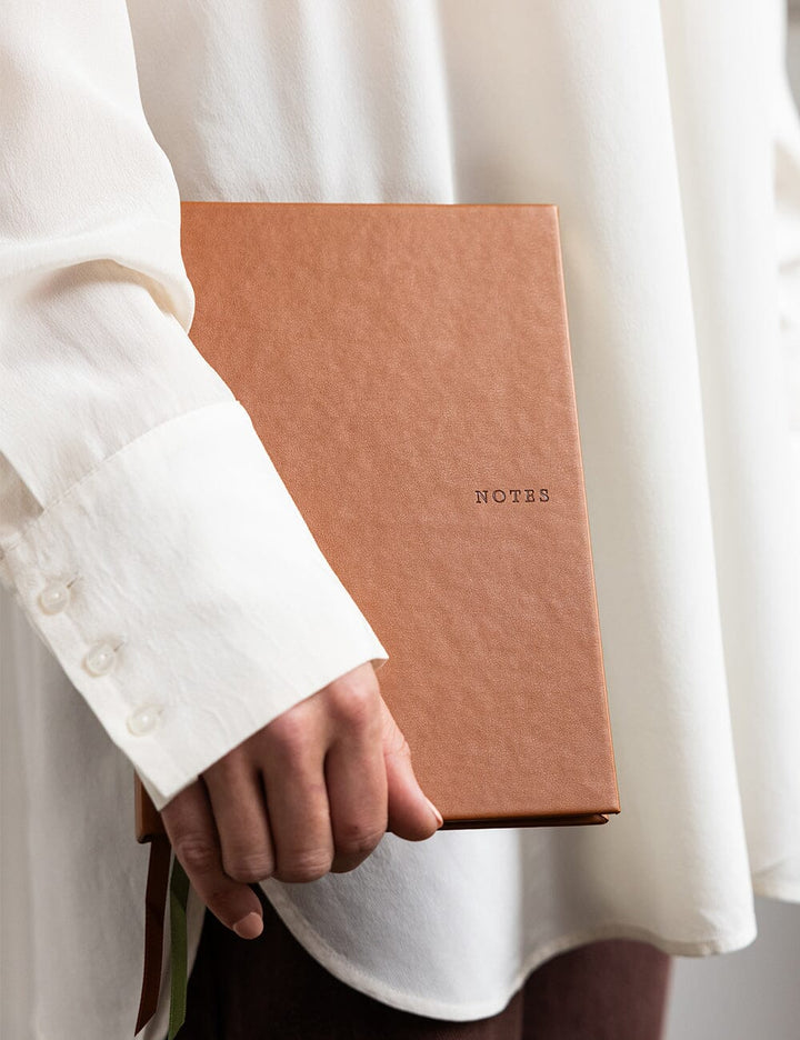 Person holding a brown notebook titled 'NOTES' with a white shirt background