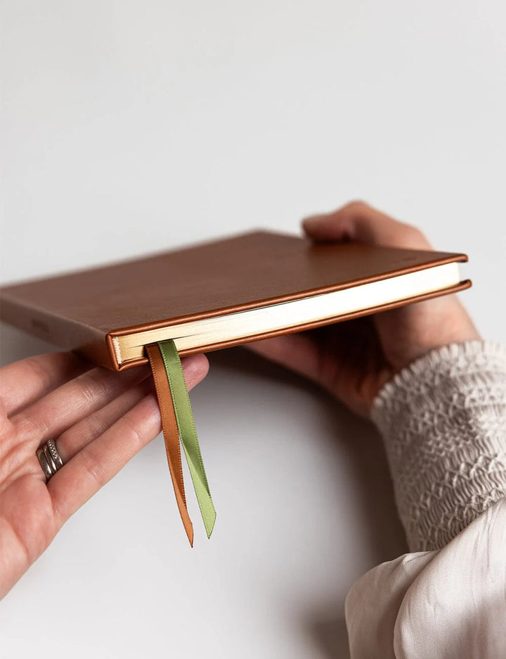 Person holding a brown notebook with a green ribbon bookmark on a light gray background