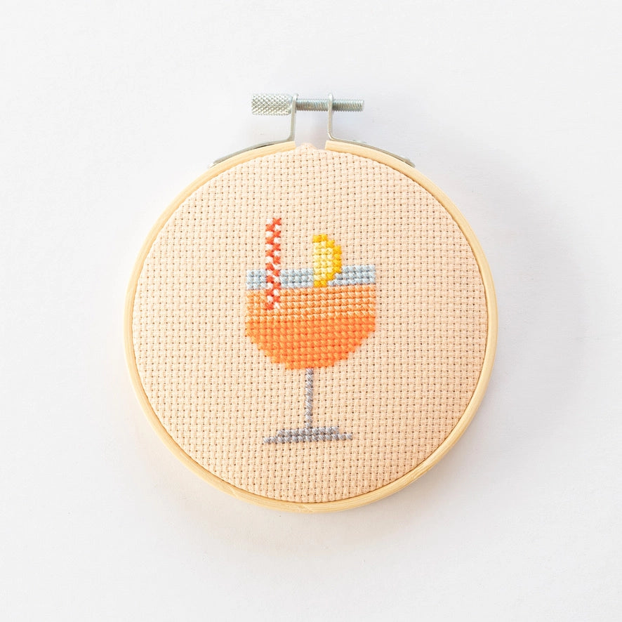 Cross-stitched design of a cocktail glass with an orange drink and lemon wedge in a wooden hoop on a white background.