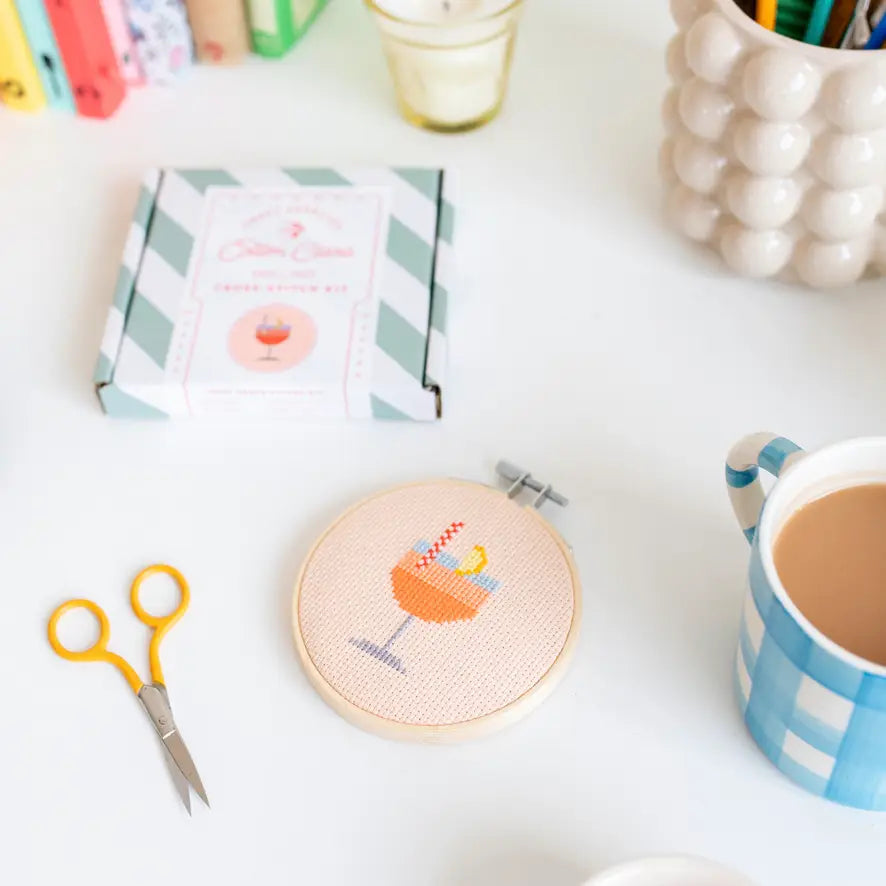 Cross-stitched hoop with a drink design on a table with scissors and a mug.