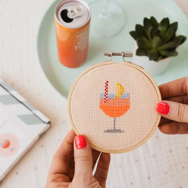 Hand holding a cross-stitched hoop with a cocktail design, with a can and glass in the background.