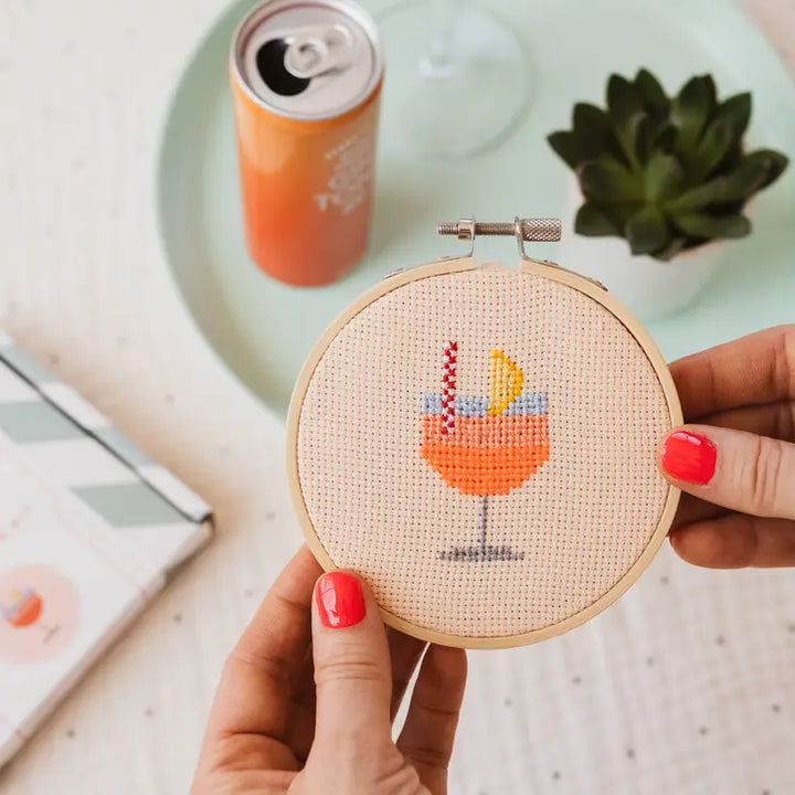 Hand holding a cross-stitched hoop with a cocktail design, with a can and glass in the background.