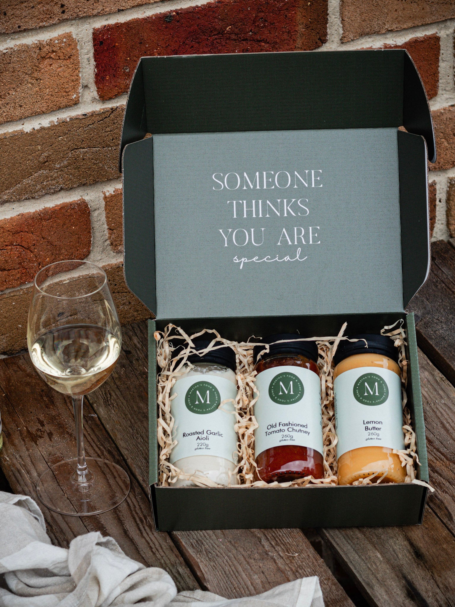 Gift set with three bottles and a glass of white wine on a wooden surface with a brick wall background.