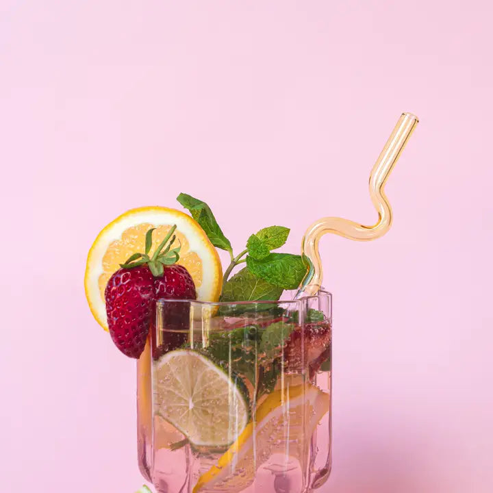 Glass with a cocktail with lemon slices, strawberry, lime, and mint on a pink background with a curly yellow glass straw. 
