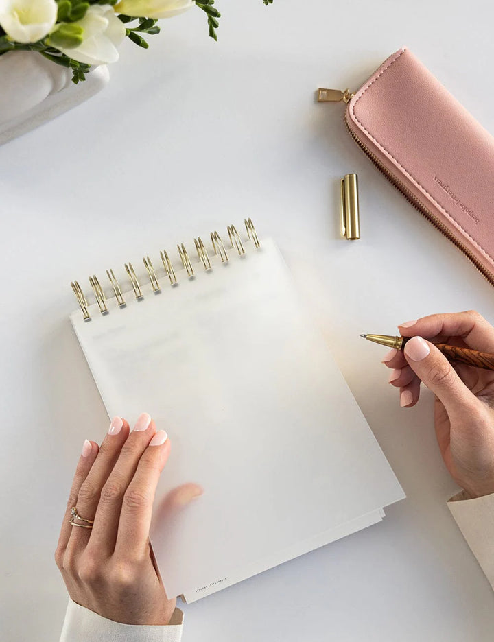 Person writing in a notebook with a pen, next to a pink wallet and flowers on a white surface
