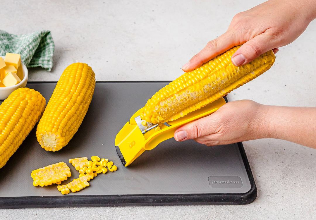 Person using a corn peeler to remove kernels from an ear of corn on a cutting board