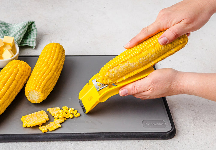 Person using a corn peeler to remove kernels from an ear of corn on a cutting board