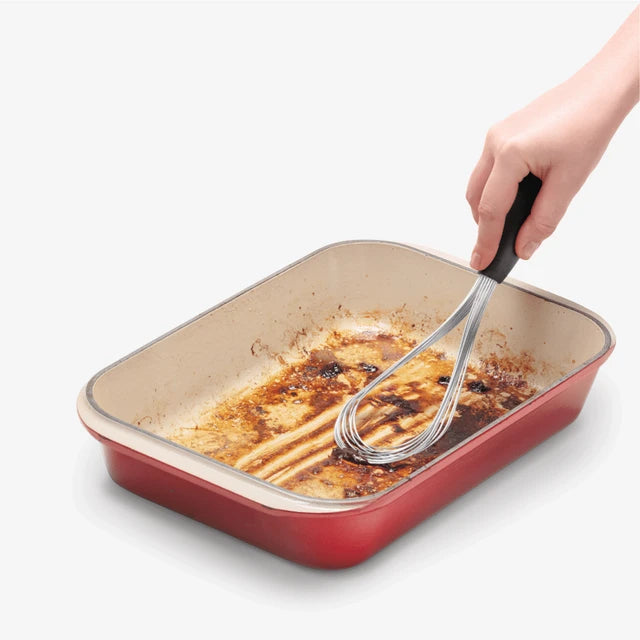 Person using a whisk to deglaze a red rectangular baking dish on a white background
