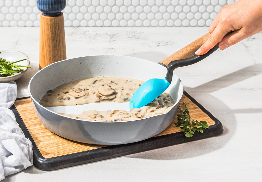 Person stirring creamy mushroom sauce in a gray pan with a blue spatula on a wooden cutting board.