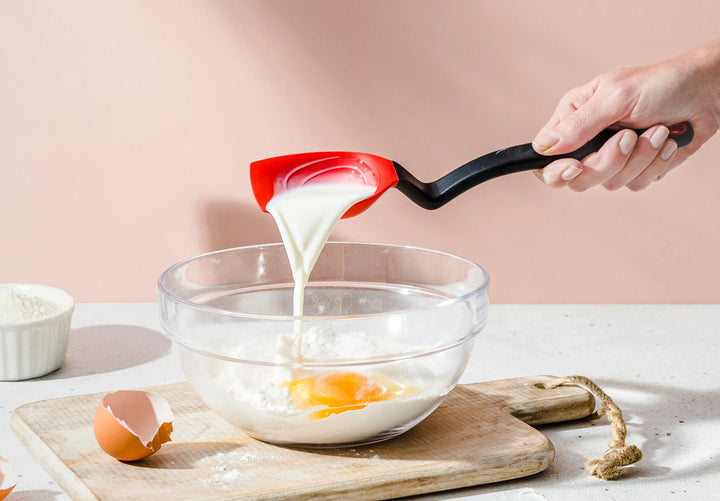 Person using a red spatula to pour liquid into a glass bowl with eggs on a light pink background