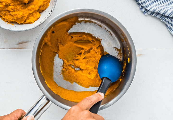 Person using a blue spoon to scoop pumpkin puree from a pot on a white surface.