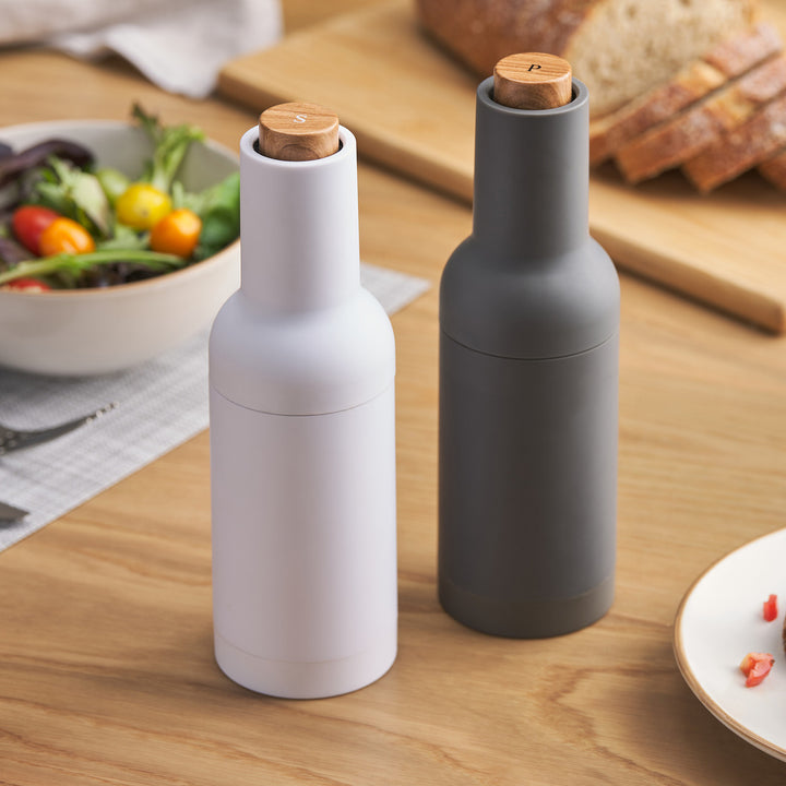 Salt and pepper mills, one white and one gray, with wooden caps on a wooden table with food items.