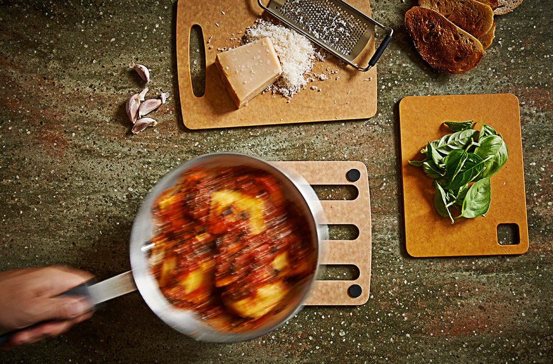 Person cooking pasta with sauce in a pan on a rustic stone surface with ingredients like cheese and basil.