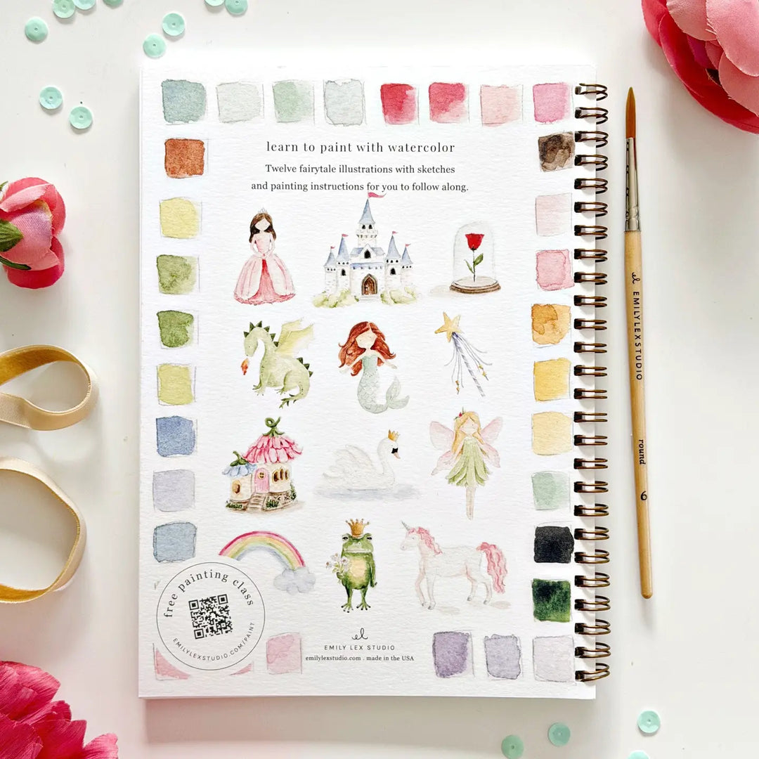 Watercolor art book with illustrations of fairies, castles, and other fantasy elements on a white surface with flowers and a paint brush.