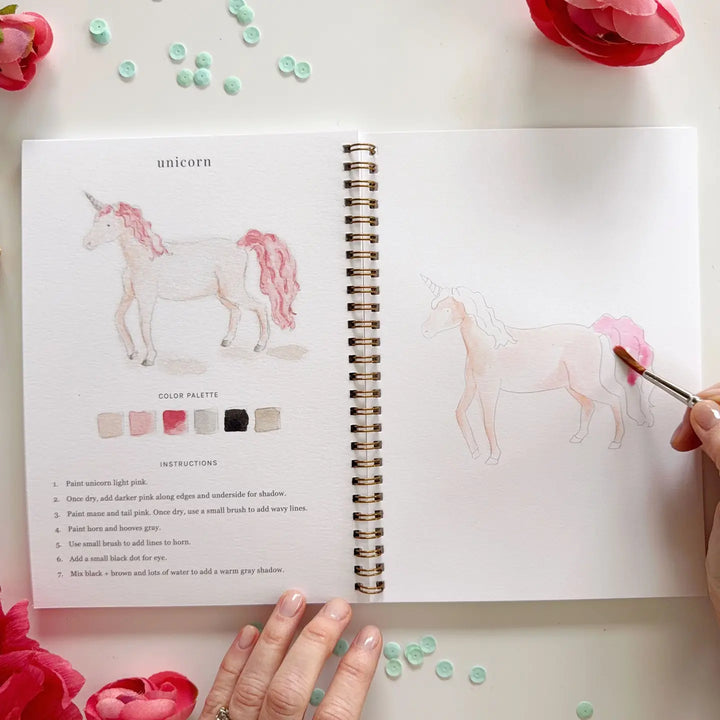 Open book with unicorn illustration and color palette, surrounded by flowers and paintbrush.