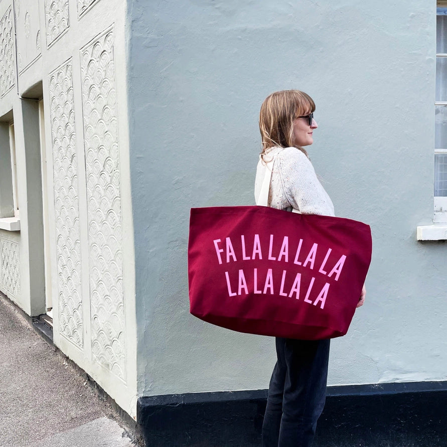 A woman stands against a pale blue-gray wall holding a large burgundy tote bag printed with the phrase &