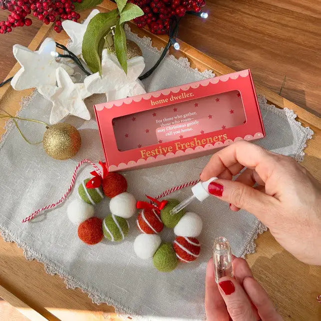 Festive freshener packaging with a hand holding a small bottle, surrounded by Christmas decorations on a wooden surface.