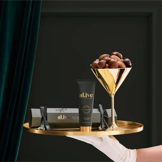 Gold tray with 'al.ive' skincare product, cocktail glass with chestnuts, and green curtain on black background