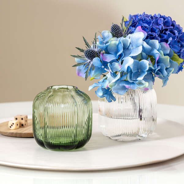 Two glass vases with ribbed texture, one green and one clear, containing blue flowers on a white surface.