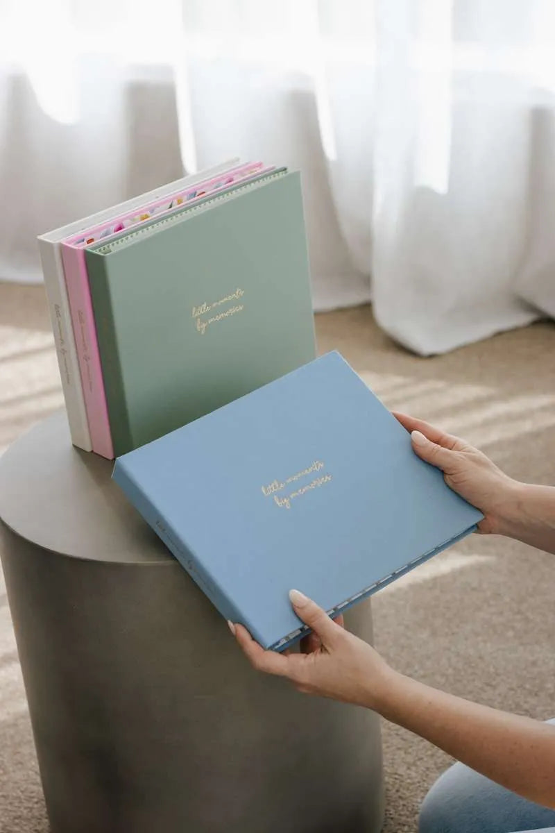 Person holding a blue photo album with other albums stacked on a table against a light curtain background.