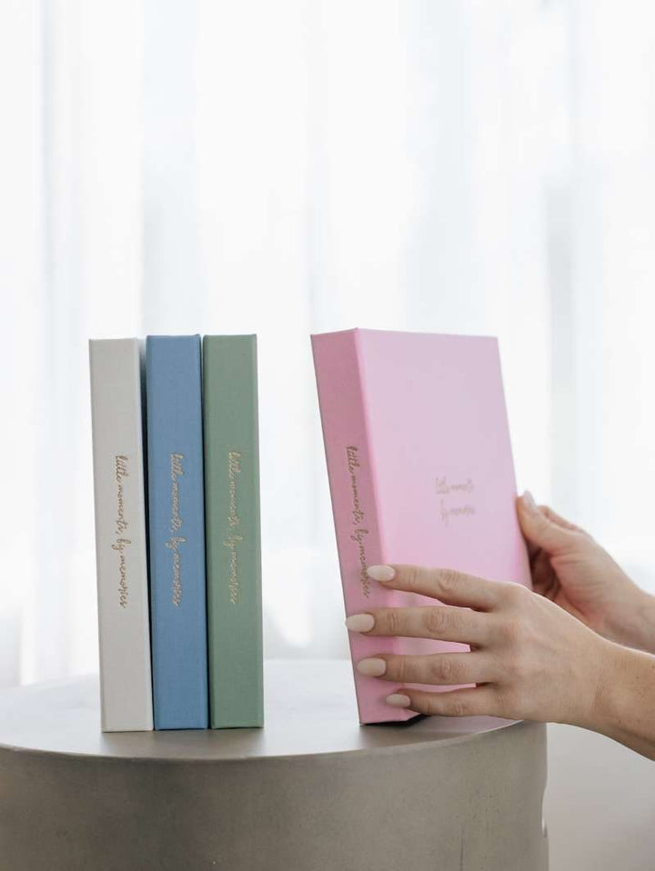 Three books of different colors on a surface with a hand reaching to pick up a pink book.