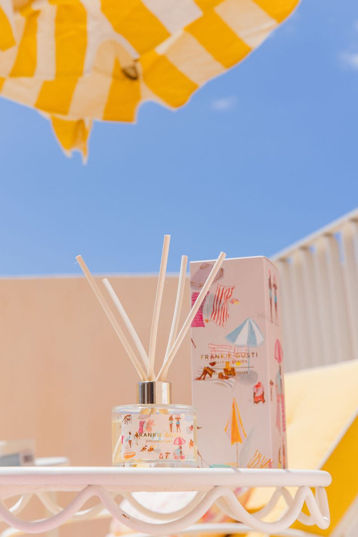 Reed diffuser and packaging on a table with a blue sky and yellow umbrella in the background