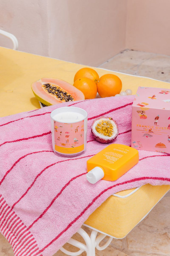 Pink towel with a Frankie Gusti candle, oranges, papaya, and a box on a yellow bed with a pink beach towel.