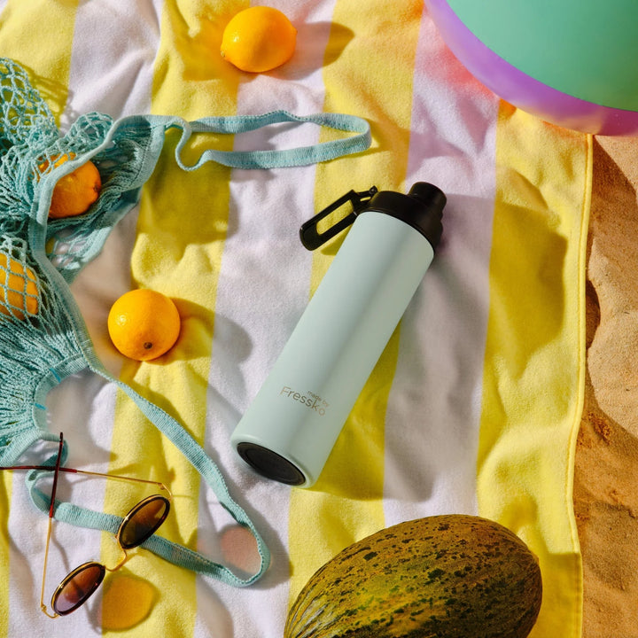 Light blue water bottle with black cap on a yellow and white striped fabric with oranges and sunglasses.