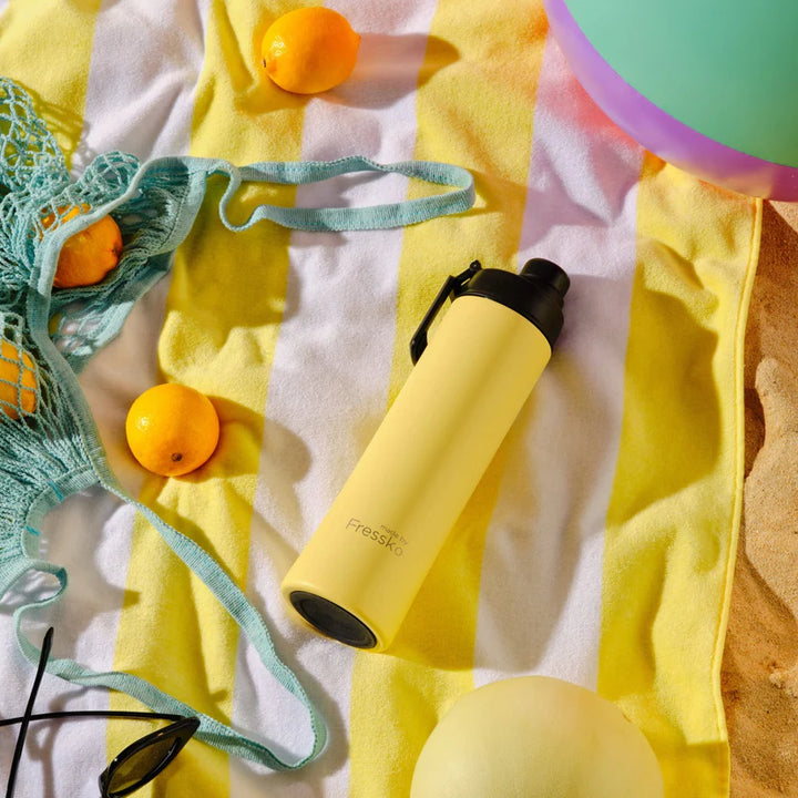 Yellow water bottle with black cap on a beach towel with sunglasses and oranges.